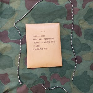 May include: A silver-toned metal ball chain necklace and a tan paper envelope printed with the text 8455-261-6629, NECKLACE, PERSONNEL, IDENTIFICATION TAG, 1 EACH, and DSA100-71-C-0521. The chain is composed of small spherical metal beads.