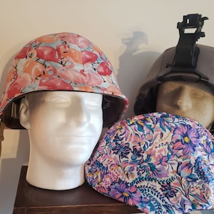 May include: Two military helmets with different fabric covers. One helmet has a pink and white flamingo print, the other has a camouflage print. Both helmets are on a wooden shelf.