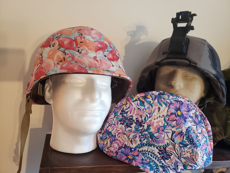 May include: Two military helmets with different fabric covers. One helmet has a pink and white flamingo print, the other has a camouflage print. Both helmets are on a wooden shelf.