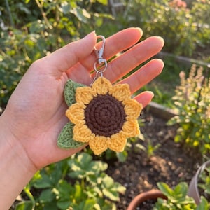 May include: A crochet sunflower keyring featuring a dark brown centre, bright yellow petals, and a single green leaf attached to a silver-toned metal lobster clasp. The sunflower is crafted with yarn and has a textured, circular design.