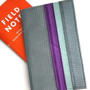 May include: Gray notebook cover with purple and teal stripes. The cover is next to an orange Field Notes notebook. The cover has a grid pattern and is stitched around the edges.