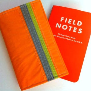 May include: An orange notebook cover with a vertical stripe of gray and green. Next to it is a red notebook with the words "FIELD NOTES" in white. The notebook cover is made of a durable material.