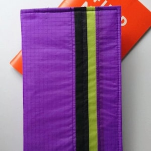 May include: A rectangular purple case with a black and lime green stripe down the center. An orange object with white text is partially visible behind the case. The case has a grid pattern.