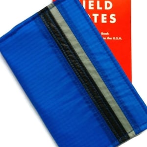 May include: A bright blue notebook cover with a black and gray stripe detail. The cover is rectangular and has a textured surface. A red notebook with the words "FIELD NOTES" is behind the cover.