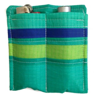 May include: A small, green pouch with two pockets. The pouch has blue and green stripes and is made of a fabric material. A lighter and a small tube are visible in the pockets.