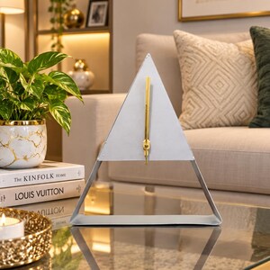 Peut inclure: Une horloge triangulaire en métal avec une finition argentée mate et une aiguille verticale de couleur dorée centrée sur le cadran. L'horloge présente un cadre géométrique minimaliste reposant sur une base plate, formant une silhouette pyramidale.