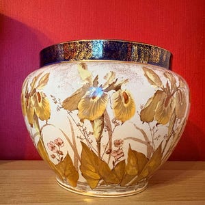 May include: A round ceramic vase featuring a cream-colored body decorated with painted yellow and brown iris flowers and green foliage. The vase has a wide, dark blue rim with a metallic gold-toned finish along the top edge.