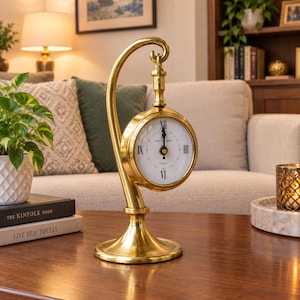 May include: A brass table clock with a curved stand and a circular face featuring black Roman numerals and the text 49 BOND STREET LONDON. The clock has a white dial with black hands and is suspended from the top of the polished brass frame.