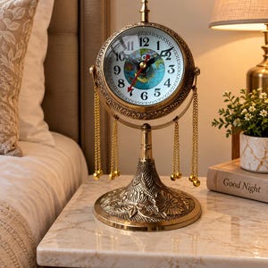 May include: A gold-toned metal table clock featuring a circular face with a world map illustration, black Arabic numerals, and red hands. The clock is mounted on a pedestal base with floral engravings and is adorned with hanging gold chains.