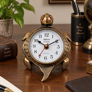 May include: A bronze-toned turtle-shaped desk clock with a white face, black numerals, and red eyes. The clock face features the text SMITH, 8 DAYS, and London. It has black hour and minute hands and a red second hand, standing on four legs.