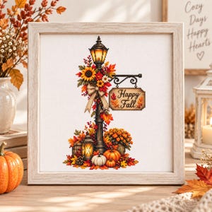 May include: A framed cross-stitch embroidery featuring a black street lamp decorated with autumn leaves, sunflowers, and a beige bow. A hanging sign reads Happy Fall. The base includes pumpkins, a glowing lantern, and fall foliage on white fabric.