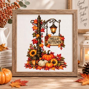 May include: A framed cross-stitch embroidery featuring a dark lamp post entwined with autumn leaves, a hanging sign reading HELLO AUTUMN, a glowing lantern, sunflowers, and orange and white pumpkins on a white fabric background.