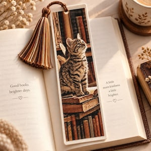 May include: A rectangular bookmark featuring a cross-stitch design of a brown tabby cat sitting on a stack of books in a library. The bookmark has a white border and a brown and tan tassel attached to the top. The design includes vertical book spines.