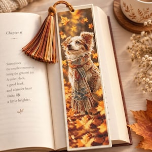 May include: A rectangular bookmark featuring a cross-stitch design of a dog wearing a patterned scarf, surrounded by falling autumn leaves. The bookmark is topped with a brown tassel and is placed inside an open book with printed text.