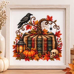 May include: A framed cross-stitch embroidery featuring a stack of books forming a pumpkin shape, topped with a black raven, autumn leaves, red berries, a glowing lantern, a small spider, and a human skull, all set against a white fabric background.