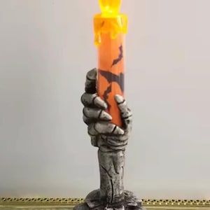 Peut inclure: Un bougeoir décoratif composé d'une main squelettique grise émergeant d'une base de crânes, tenant une bougie pilier orange ornée de silhouettes de chauves-souris noires et surmontée d'une flamme LED jaune avec des coulures de cire.