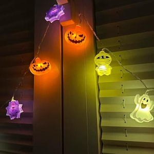 Peut inclure: Une guirlande lumineuse LED sur le thème d'Halloween composée de figurines translucides, dont des toiles d'araignée violettes, des citrouilles orange, une momie blanche et un fantôme blanc. Les lumières sont allumées et reliées par un fil fin.