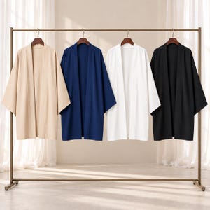Puede incluir: Cuatro kimonos abiertos colgados en un perchero de metal. Las prendas son de colores sólidos: beige, azul marino, blanco y negro. Cada kimono presenta mangas anchas de tres cuartos, un dobladillo recto y un diseño de cuello sencillo.