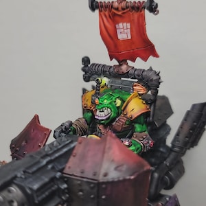 May include: A miniature green-skinned orc figure with yellow shoulder armour, riding a dark metallic vehicle with a large red banner featuring a white fist icon. The orc has a bionic eye, bared teeth, and is positioned behind a large red front plate.