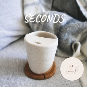 May include: A speckled white ceramic travel mug with a wooden coaster. The mug has a lid with a small opening. The word "SECONDS" is written in white across the top. A circular sticker reads "50 TREES PLANTED with your order."