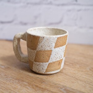 May include: A handmade ceramic mug with a geometric checkerboard pattern in tan and white. The mug has a sturdy handle and a speckled finish, perfect for coffee or tea. The mug is approximately 3.5 inches tall.