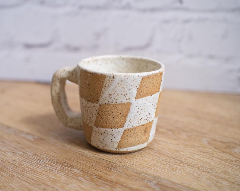 May include: A handmade ceramic mug with a geometric checkerboard pattern in tan and white. The mug has a sturdy handle and a speckled finish, perfect for coffee or tea. The mug is approximately 3.5 inches tall.