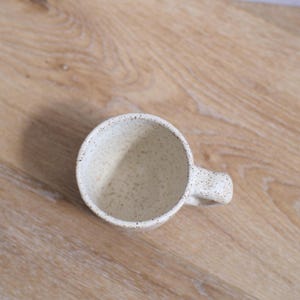 May include: A speckled off-white ceramic mug with a rounded handle. The mug has a wide opening and a slightly textured surface. The interior is a lighter shade of off-white. The mug is set against a light brown wooden surface.