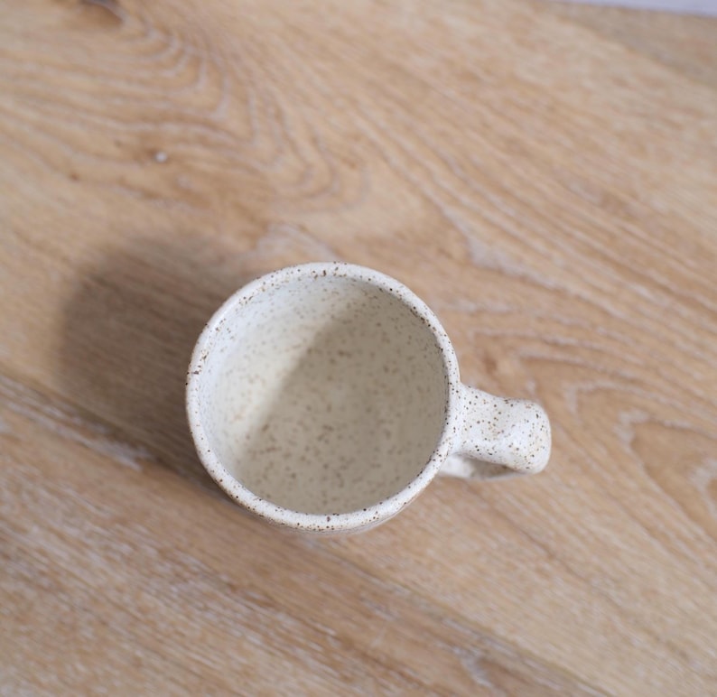 May include: A speckled off-white ceramic mug with a rounded handle. The mug has a wide opening and a slightly textured surface. The interior is a lighter shade of off-white. The mug is set against a light brown wooden surface.