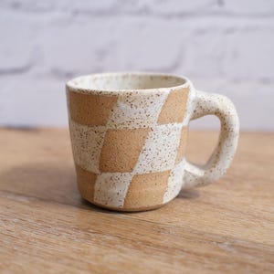 May include: A ceramic mug with a checkerboard pattern in beige and white. The mug has a speckled finish and a rounded handle. The mug is sitting on a wooden surface.