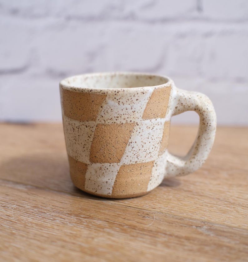 May include: A ceramic mug with a checkerboard pattern in beige and white. The mug has a speckled finish and a rounded handle. The mug is sitting on a wooden surface.