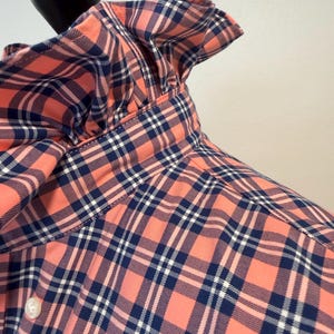 May include: A long-sleeved shirt featuring a coral, navy blue, and white plaid pattern. The shirt has a high, ruffled collar and a button-down front with a single visible white button. The fabric has a structured appearance with visible seams.