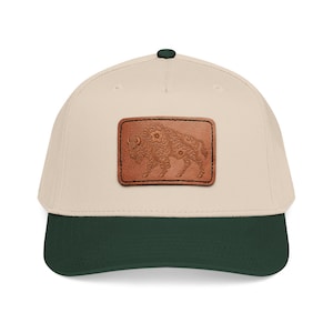 May include: A beige baseball cap with a dark green brim and a rectangular brown leather patch centered on the front. The patch features an embossed, intricate floral pattern shaped like a bison, stitched onto the cap with black thread.