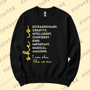 May include: A black crewneck sweatshirt featuring white text that reads: She is EXTRAORDINARY. CREATIVE. INTELLIGENT. CONFIDENT. KIND. IMPORTANT. MAGICAL. AMAZING. I am she She is me. The words She is and She is me are written in yellow script.