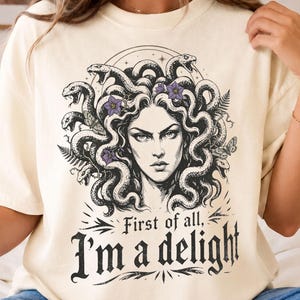 Könnte beinhalten: Ein elfenbeinfarbenes Kurzarm-T-Shirt mit einer schwarz-weißen Grafik von Medusa mit Schlangen als Haar und kleinen violetten Blumen. Unter der Illustration steht der Text First of all, I'm a delight in einer fetten, dekorativen schwarzen Schrift.