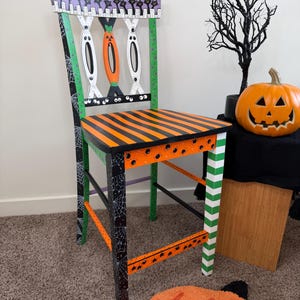 May include: A wooden chair painted with Halloween motifs. The seat features orange and black stripes. The backrest displays black cats on a purple background, with white and orange ghost-like shapes. The legs have spiderweb and stripe patterns.