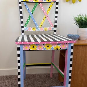 Puede incluir: Una silla de madera con asiento y detalles de marco a rayas blancas y negras. El respaldo y el faldón delantero muestran un patrón floral con flores amarillas y rosas. El respaldo tiene un diseño entrecruzado en azul, rosa y amarillo.