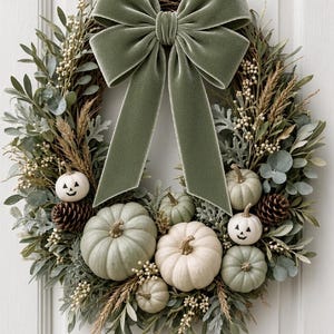 May include: A decorative wreath featuring a large sage green velvet bow at the top, accented with assorted light green and white pumpkins, two of which have painted jack-o'-lantern faces, brown pinecones, dried wheat stalks, and green foliage.