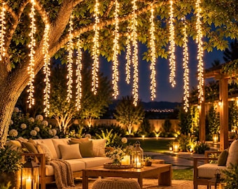 May include: Multiple strands of warm white LED fairy lights hang vertically from tree branches, creating a glowing curtain effect. The lights feature small, teardrop-shaped bulbs that emit a soft, golden illumination over an outdoor patio area.