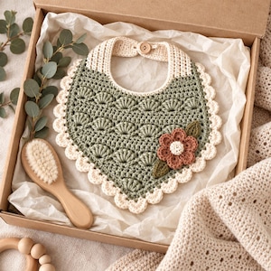 Puede incluir: Un babero de ganchillo color verde salvia con un borde festoneado color crema y un cierre de botón de madera. El babero presenta un patrón de punto de concha texturizado y está decorado con una pequeña flor de ganchillo terracota y dos hojas verdes.