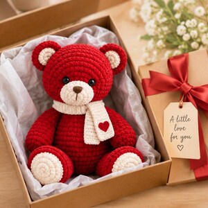 Puede incluir: Un oso de peluche tejido a ganchillo de color rojo con hocico, interior de las orejas y bufanda de color crema. La bufanda tiene un pequeño corazón rojo. El oso está en una caja de regalo de cartón con papel seda y una etiqueta A little love for you.