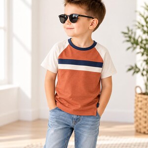 Puede incluir: Una camiseta de manga corta raglán con el torso de color naranja óxido, una franja horizontal azul marino y blanca en el pecho, mangas de color crema y cuello redondo azul marino, combinada con vaqueros de mezclilla azul claro y gafas de sol negras.