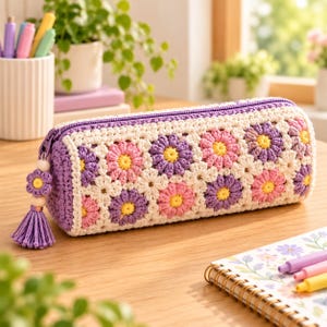 May include: A cylindrical pencil case made of crocheted yarn featuring a repeating pattern of white, pink, and purple daisy-like flowers. The case has a purple zip closure and a decorative purple tassel pull with a small matching flower charm.