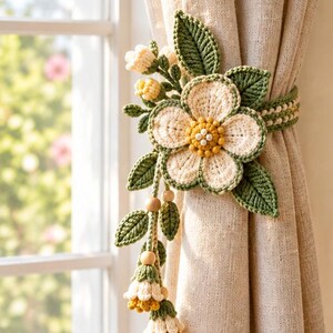 May include: A crochet curtain tieback featuring a large cream-colored flower with a yellow center, surrounded by green crocheted leaves and smaller buds. The design includes dangling floral accents with wooden beads attached to a textured fabric curtain.