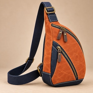 May include: A teardrop-shaped sling backpack featuring a combination of burnt orange and navy blue fabric. The bag includes two front zippered compartments with brown leather-like pull tabs, antique brass hardware, and an adjustable navy blue strap.