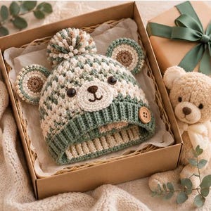 May include: A crochet beanie featuring a bear face design with round ears, a pom-pom on top, and a ribbed brim in sage green and cream. The hat has dark brown button eyes, a stitched nose and mouth, and a single wooden button on the side.