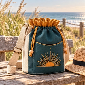 Può includere: Una borsa a secchiello in tela color verde acqua e beige con chiusura a coulisse, caratterizzata da un ricamo a raggiera giallo senape sulla tasca frontale. La borsa include una tracolla beige regolabile e cordini giallo senape con perline.