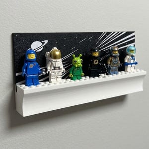 May include: A white wall-mounted display shelf featuring a black back panel with a white space-themed graphic of stars and a planet. Six small plastic astronaut and alien figures are standing on the white studded surface of the shelf.