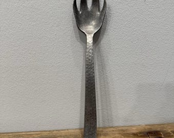 Könnte beinhalten: Eine silberfarbene Salatgabel aus Metall mit einem langen, geraden Griff und einem breiten, dreizinkigen Kopf. Die gesamte Oberfläche des Bestecks weist eine markante Hammerschlagstruktur auf, die dem Metall ein mattes Aussehen verleiht.
