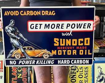 Puede incluir: Letrero metálico vintage de Sunoco Motor Oil con un hombre musculoso tirando de un coche clásico. El letrero tiene un fondo azul marino con texto blanco y amarillo. El texto dice "Avoid Carbon Drag" y "Get More Power".
