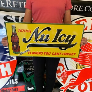 May include: A vintage metal sign advertising Nu Icy, a soda brand, with the slogan "Drink Nu Icy Assorted Flavors You Can't Forget". The sign features a yellow background with green and red accents, and a bottle of soda with a red label.